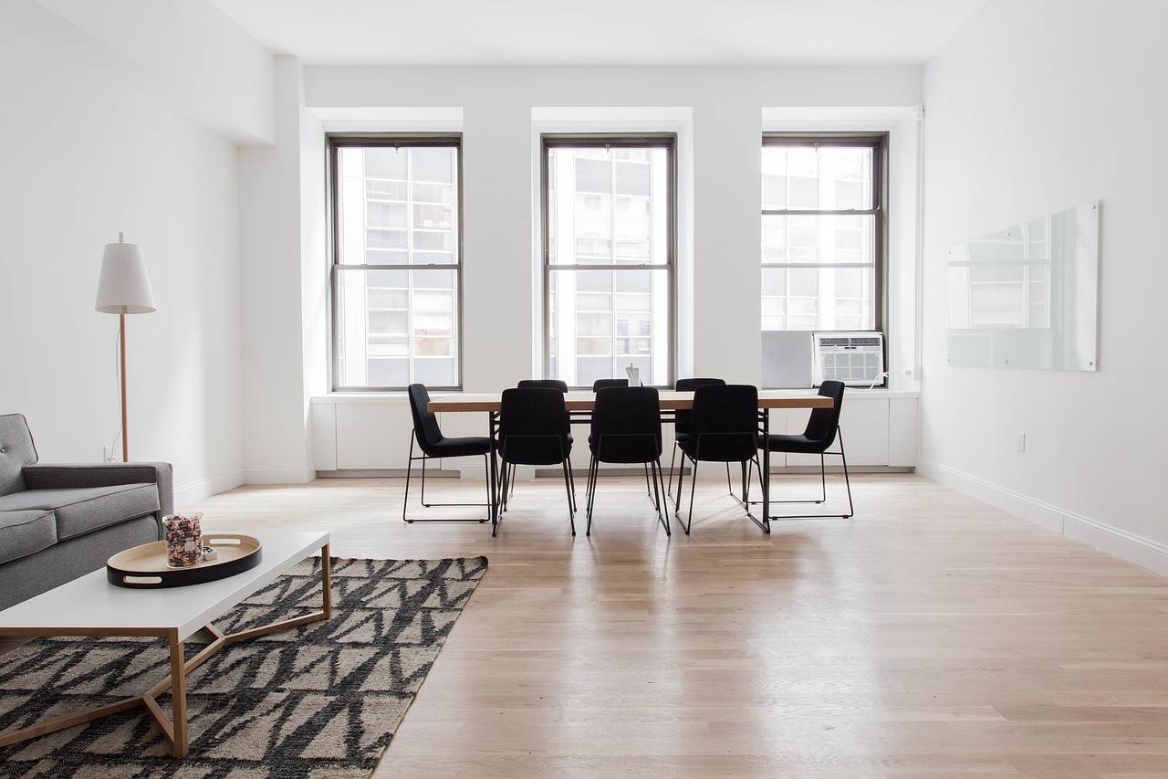 Zur Vermietung steht ein minimalistischer Wohn- und Essbereich mit hellem Holzboden zur Verfügung. Er verfügt über ein graues Sofa, einen geometrischen Teppich, einen modernen Couchtisch und einen Esstisch mit schwarzen Stühlen. Drei große Fenster lassen Tageslicht herein und verleihen dem Raum Helligkeit.