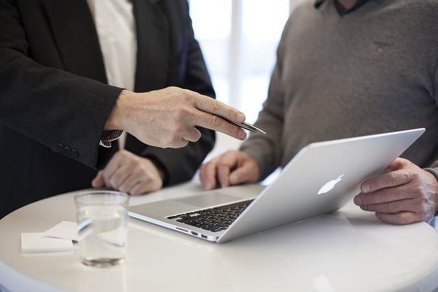 Zwei Personen stehen an einem runden Tisch, konzentrieren sich auf einen Laptop und sind in Vermittlung verwickelt. Eine Person zeigt mit einem Stift auf den Bildschirm, während die andere zusieht. Auf dem Tisch daneben stehen ein Glas Wasser und einige Papiere.