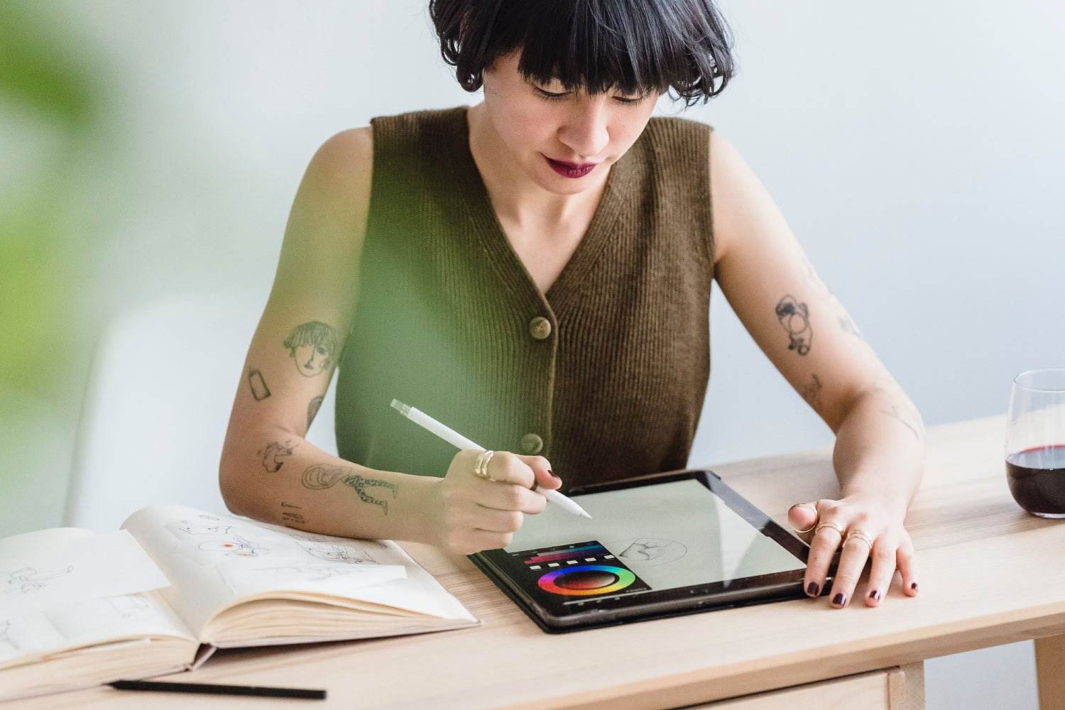 Ein tätowierter Künstler zeichnet mit einem Stift auf einem Tablet, daneben liegt ein offenes Skizzenbuch. Sie sitzen an einem Holztisch mit einem Glas Rotwein neben sich und die Umgebung strahlt helle Kreativität aus. Dieser Moment fängt die Vermittlung von Inspiration und Kunstfertigkeit in perfekter Harmonie ein.