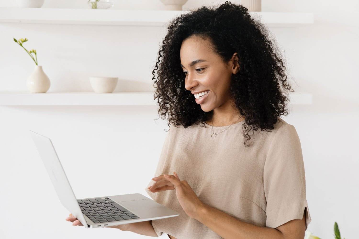 Eine Frau mit lockigem Haar lächelt, während sie auf einen Laptop blickt, den sie in der Hand hält, und möglicherweise gerade ein Vermittlungsdokument durchsieht. Sie trägt ein beiges Oberteil und steht in einem hellen Raum mit minimalistischen Regalen, die mit kleinen Pflanzen und Vasen dekoriert sind.