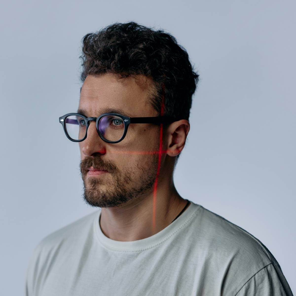 Ein Mann mit lockigem Haar und Brille, möglicherweise von der Vires-Gruppe, blickt zur Seite und trägt ein helles Hemd. Auf seinem Gesicht und Hemd werden vor einem schlichten Hintergrund rote Laserlinien projiziert.