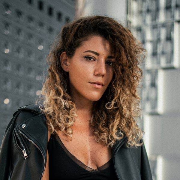 Eine Person mit lockigem Haar, die eine schwarze Lederjacke und ein schwarzes Lederoberteil trägt, steht selbstbewusst in einer städtischen Umgebung mit modernen Gebäuden im Hintergrund und verkörpert den mutigen Geist der Vires Group.