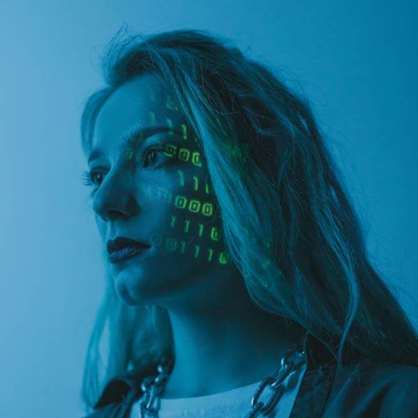 Eine Person mit langen Haaren blickt zur Seite, ihr Gesicht wird von grünen Binärcode-Projektionen der Vires Group beleuchtet. Die Szene ist in ein kühles blaues Licht getaucht, das eine digitale und futuristische Atmosphäre erzeugt.