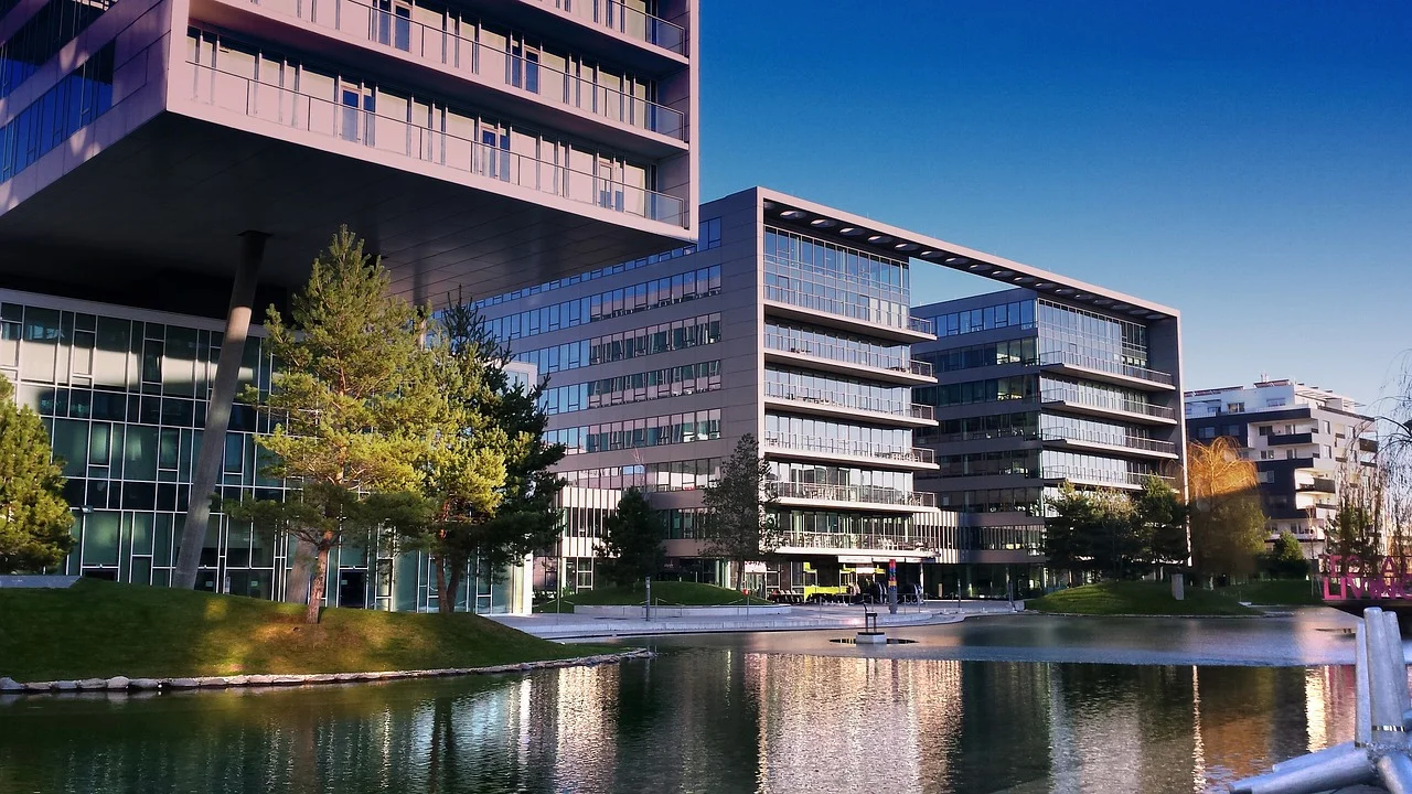 Ein moderner Bürokomplex, der Hauptsitz der Vires Group, verfügt über große Glasfenster mit Blick auf einen ruhigen Teich, in dem sich die eleganten Gebäude spiegeln. Die umliegenden grünen Bäume und der klare blaue Himmel werten die zeitgenössische Architektur zusätzlich auf.