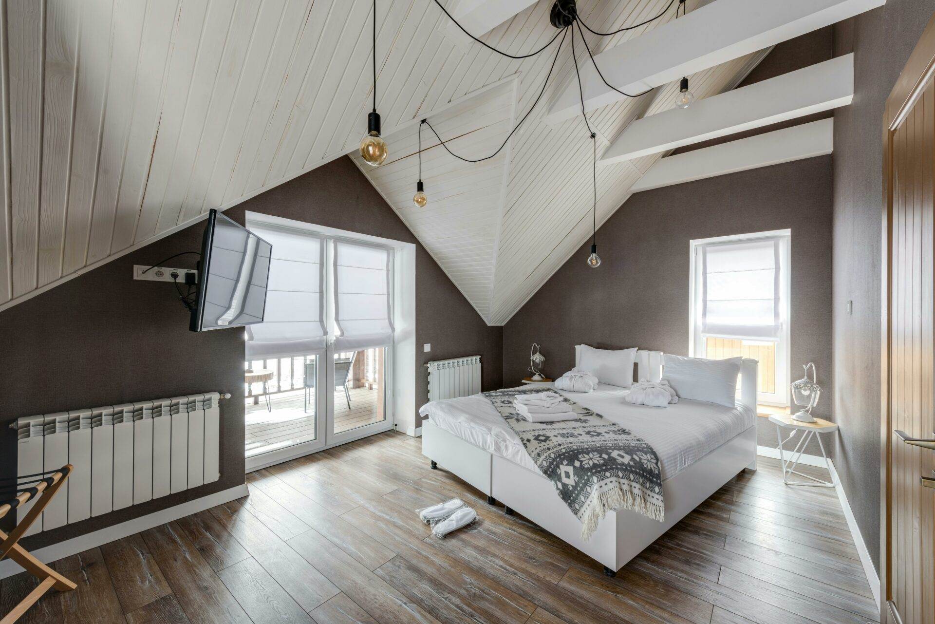 Ein gemütliches Schlafzimmer mit schrägen Holzdecken und sanfter Beleuchtung. Es gibt ein Doppelbett mit weißer Bettwäsche und einer gemusterten Decke. Ein Flachbildfernseher ist an der Wand montiert und große Fenster bieten einen Blick nach draußen. Ideal für diejenigen, die ihre Immobilie über Vires Immobilienmakler in Wien verkaufen möchten.