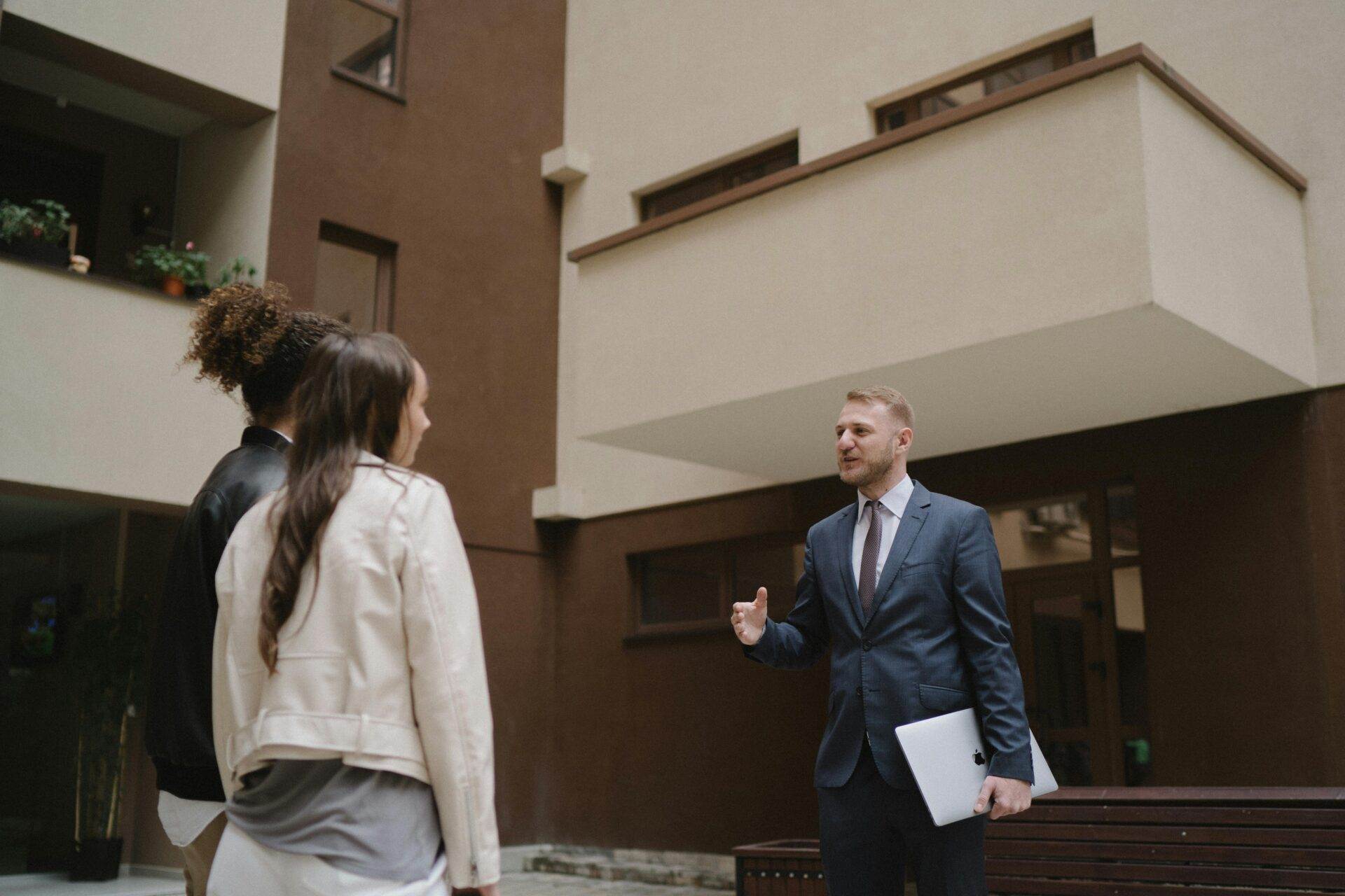 Ein Mann im Anzug mit einem Laptop in der Hand gestikuliert, während er mit zwei Personen über Immobilienkauf bei Makler spricht. Sie stehen im Innenhof eines modernen Gebäudes, im Hintergrund sind braune und beige Wände zu sehen.