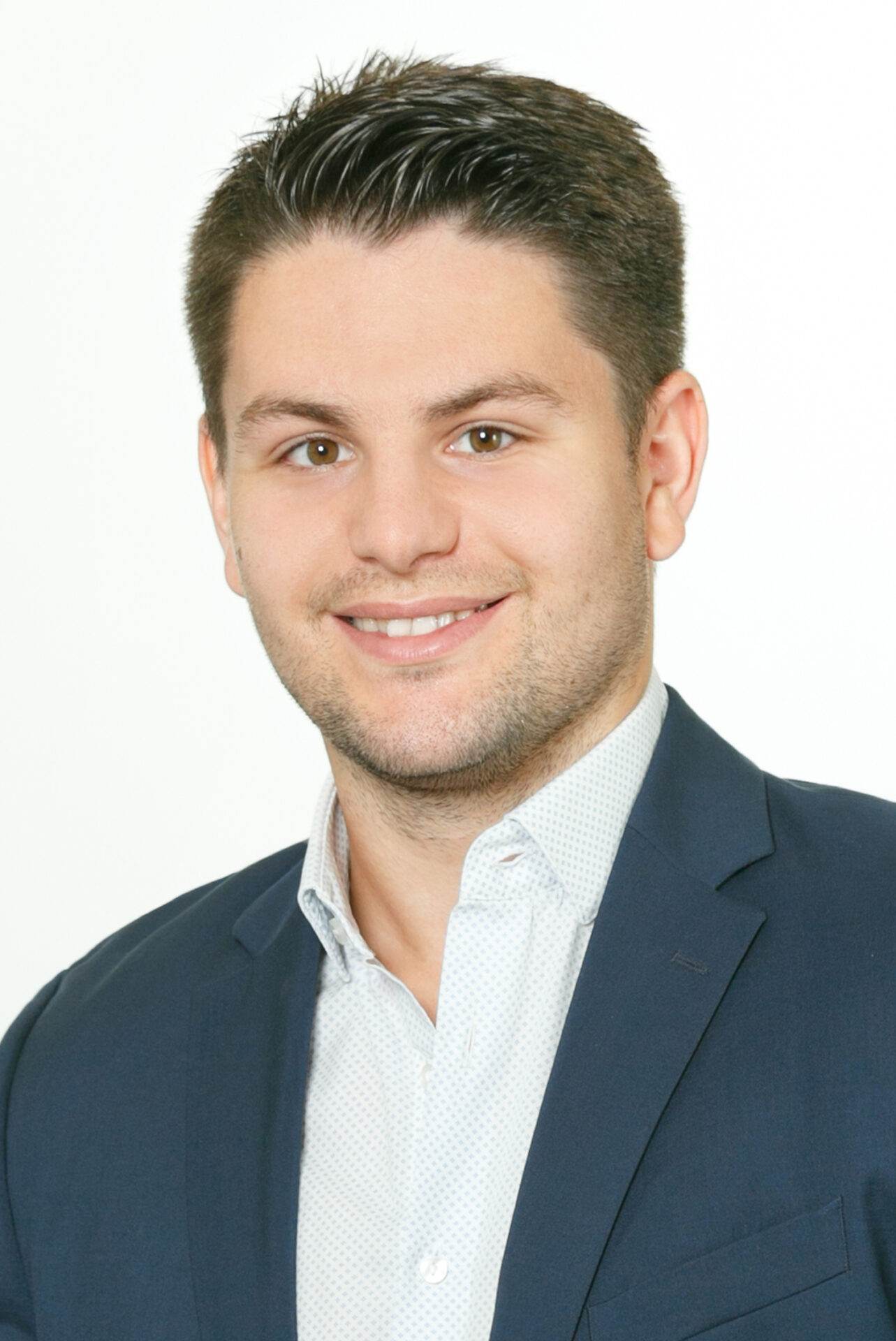 Ein Mann mit kurzen dunklen Haaren und hellem Bart lächelt in die Kamera. Er trägt ein weißes Hemd und einen dunklen Blazer. Der Hintergrund ist schlicht weiß.