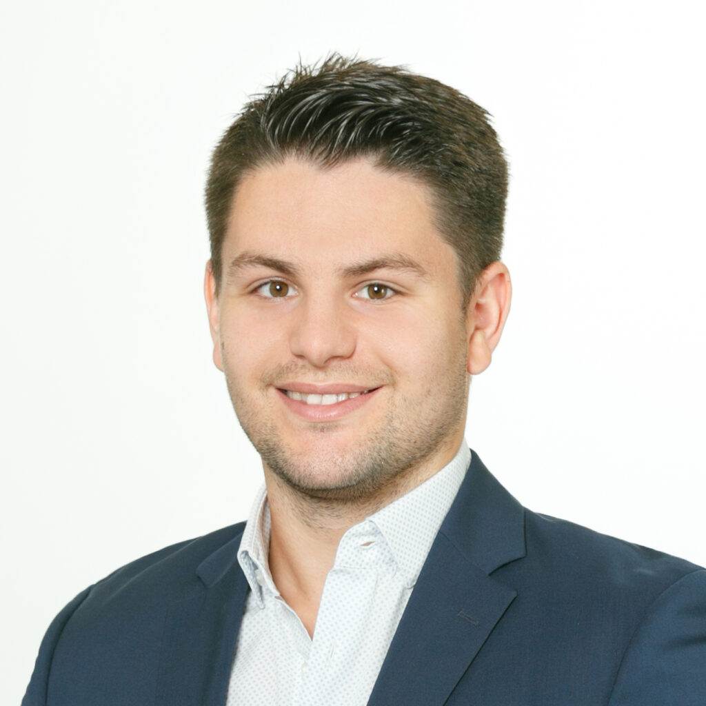 Ein Mann mit kurzen, dunklen Haaren lächelt. Er trägt einen dunklen Blazer über einem hellen Hemd. Der Hintergrund ist schlicht weiß.