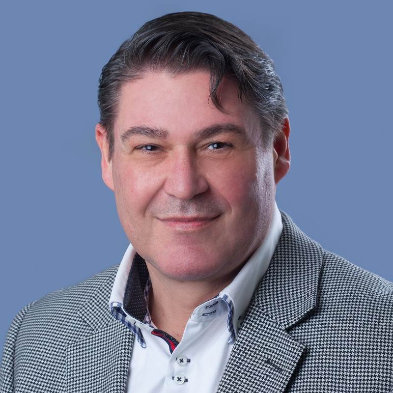 Porträt einer Person mit kurzen dunklen Haaren, die einen graukarierten Blazer und ein weißes Hemd mit Kragen trägt und sanft vor einem blauen Hintergrund lächelt.