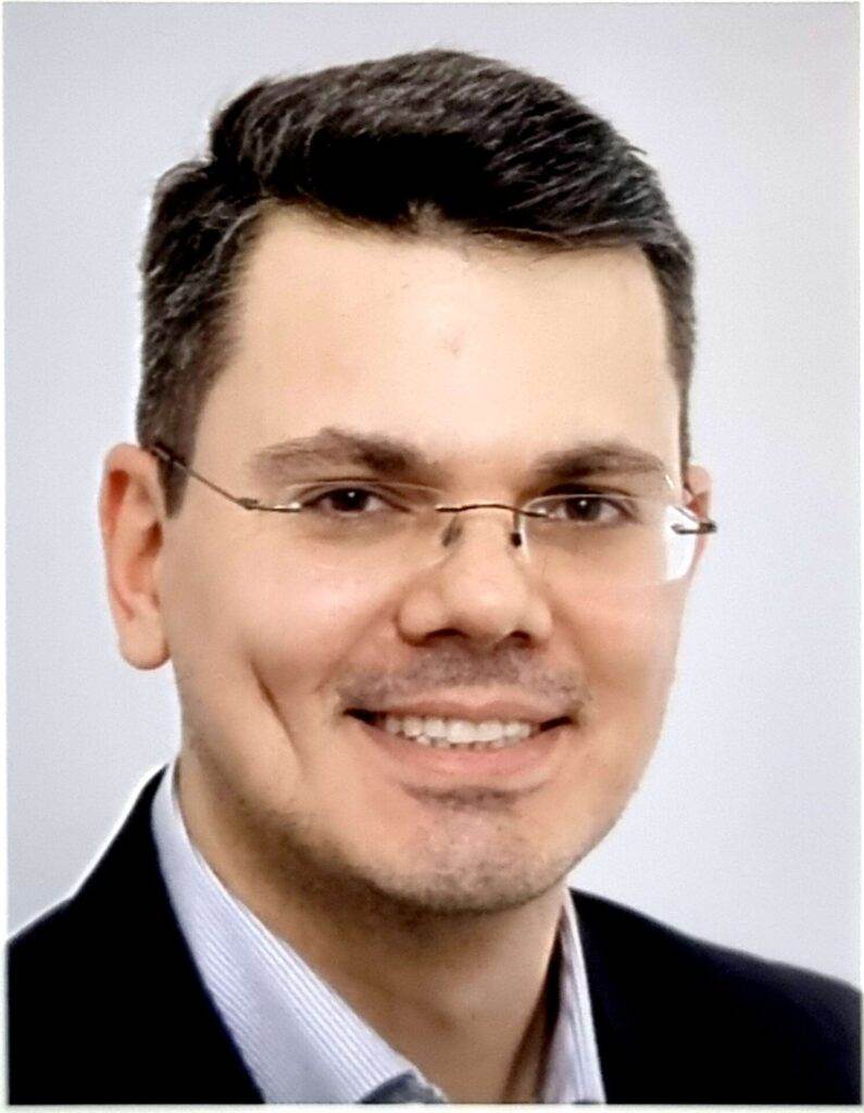 Ein Mann mit kurzen dunklen Haaren und Brille lächelt in die Kamera. Er trägt ein dunkles Jackett und ein helles Hemd. Der Hintergrund ist neutral und konzentriert sich auf sein Gesicht.