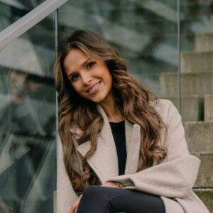 Frau mit langem welligem Haar und beigem Mantel sitzt auf Stufen neben einem Glasgeländer und lächelt in die Kamera. Im Hintergrund ist eine verschwommene Außenkulisse zu sehen.