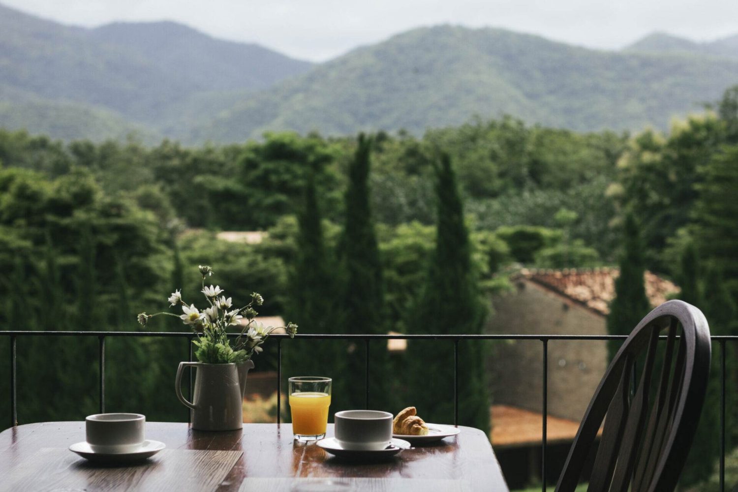 Auf einem Tisch auf einem Balkon sind die wichtigsten Utensilien für eine gemeinsame Veranstaltung ausgestellt: zwei Tassen, ein Glas Orangensaft, eine Vase mit Blumen und ein Teller mit Gebäck. Im Hintergrund sind üppige grüne Bäume und sanfte Berge unter einem bewölkten Himmel zu sehen.